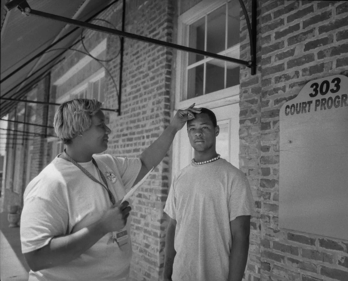 Black and white portrait taken outside of brick building of a young man and his mother, who has her hand on his head, and is smiling